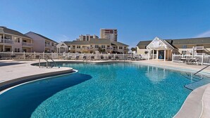 A swimming pool with a white fence, a covered seating area, and a building with balconies in the background. - Pacifico Destin SPC 2135! An exquisite condo experience right on the beach! (Destin)