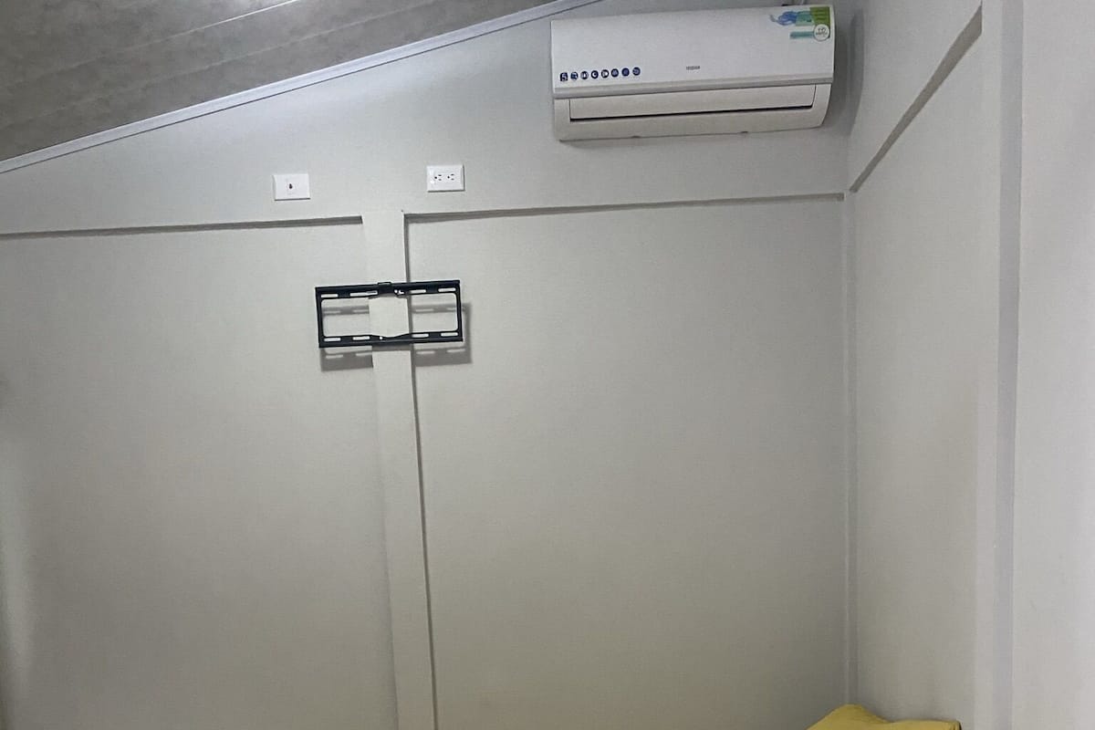 A hotel room with a bed, a white bedside table, a wooden nightstand, a remote control, and an air conditioning unit on the wall.