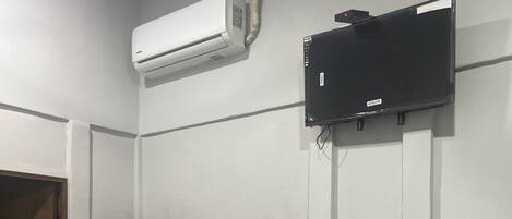 Una habitación con una cama individual alta y baja, un escritorio con computadora, aire acondicionado y un televisor montado en la pared.