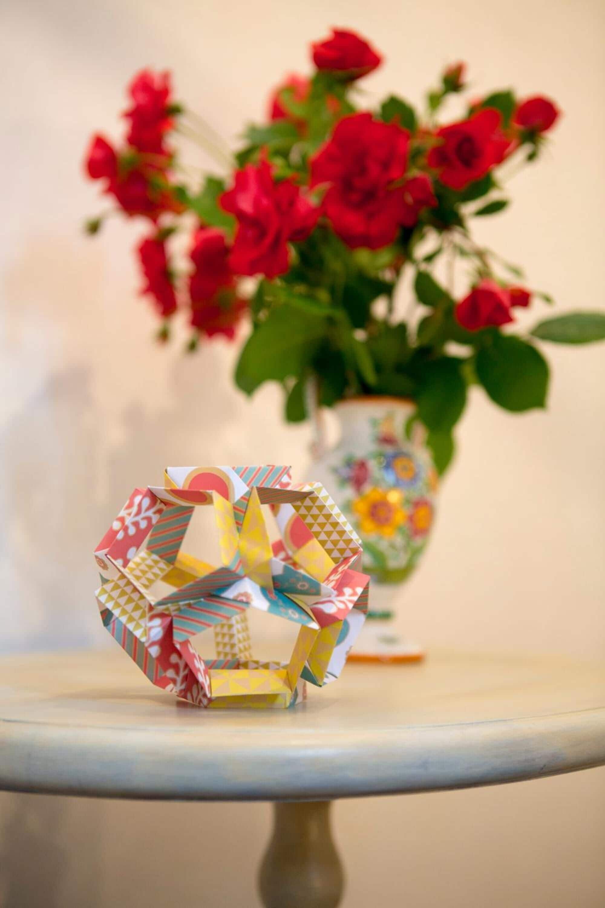 A table with a colorful origami structure and a vase of red flowers in the background.