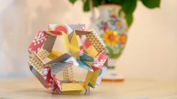 A table with a colorful origami structure and a vase of red flowers in the background.