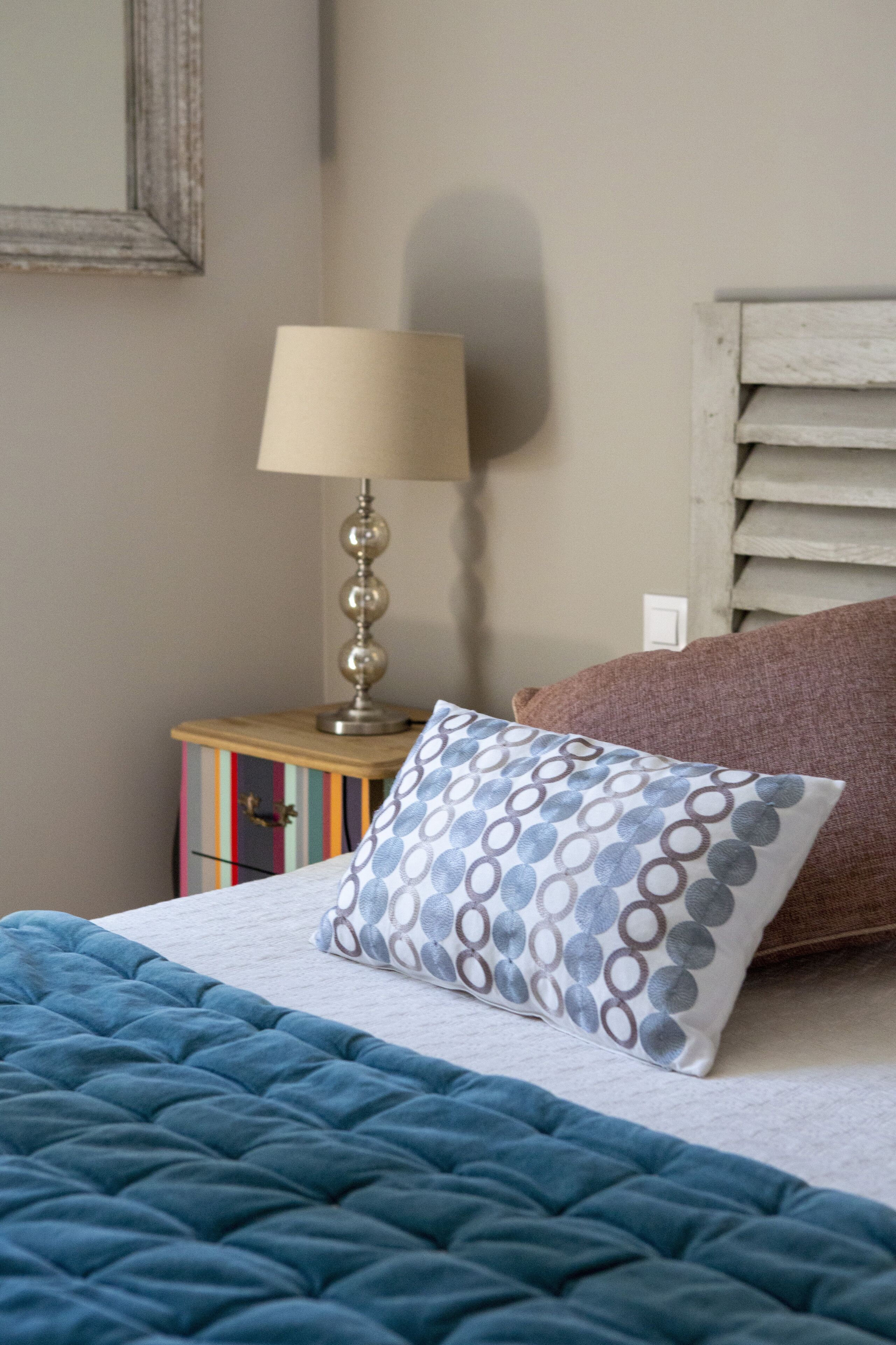 Un lit bien fait, recouvert d’une couette bleue, avec un oreiller à motifs, une lampe de chevet et une petite table de nuit en bois surmontée d’une commode à rayures colorées.