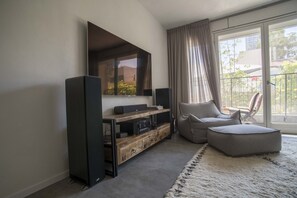 Een moderne woonkamer met een flatscreen televisie, een houten televisiemeubel, een grijze bank en een groot raam met gordijnen.