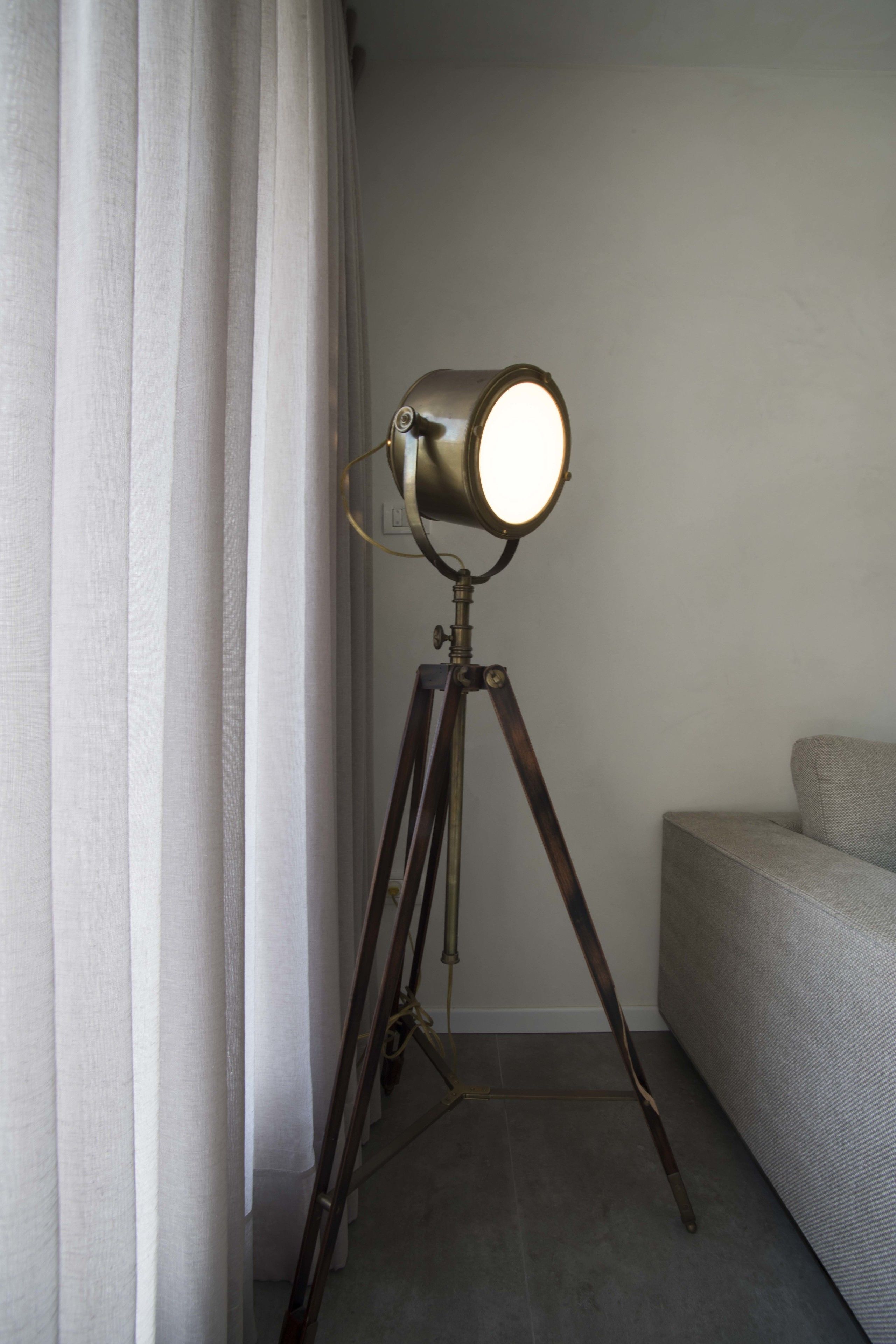 A floor lamp with a round shade on a tripod stand, positioned next to a window with white curtains.