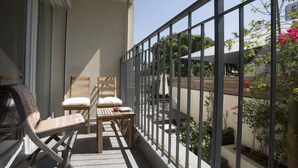 A balcony with a wooden table and two chairs, a window, and a view of a garden with potted plants and a tree. - Sasson & Co Luxury apartment Neve Tsedek (Tel Aviv)