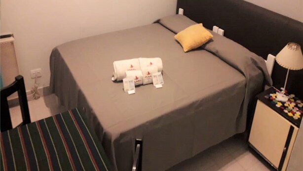 A single bed with a grey bedspread and a yellow pillow, a bedside table with a lamp, and a striped chair. - Hotel La Barca (Buenos Aires)