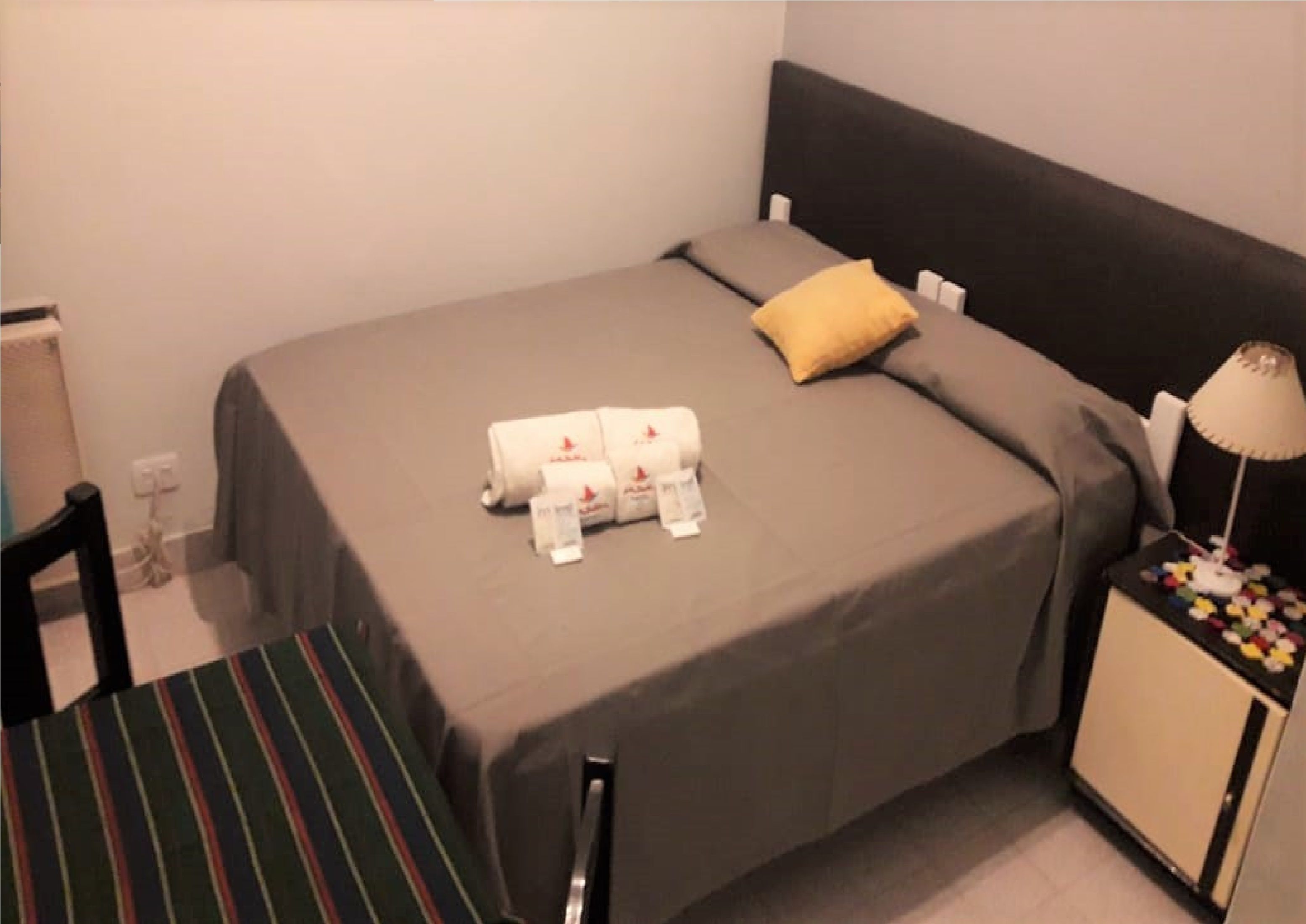 A single bed with a grey bedspread and a yellow pillow, a bedside table with a lamp, and a striped chair. - Hotel La Barca (Buenos Aires)