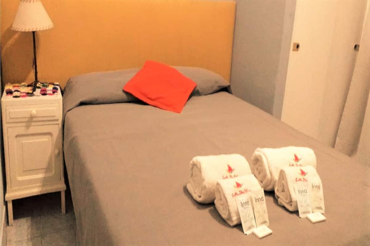 Habitación de hotel con una cama, una mesita de noche con lámpara y dos toallas enrolladas con etiquetas.