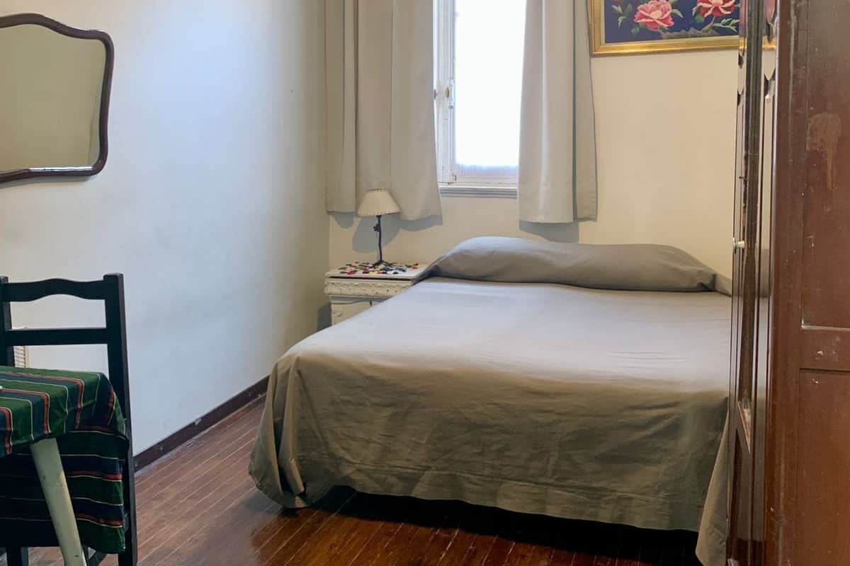 Un dormitorio con cama, una silla, una mesita de noche, ventilador de techo, ventana con cortinas y un cuadro en la pared.