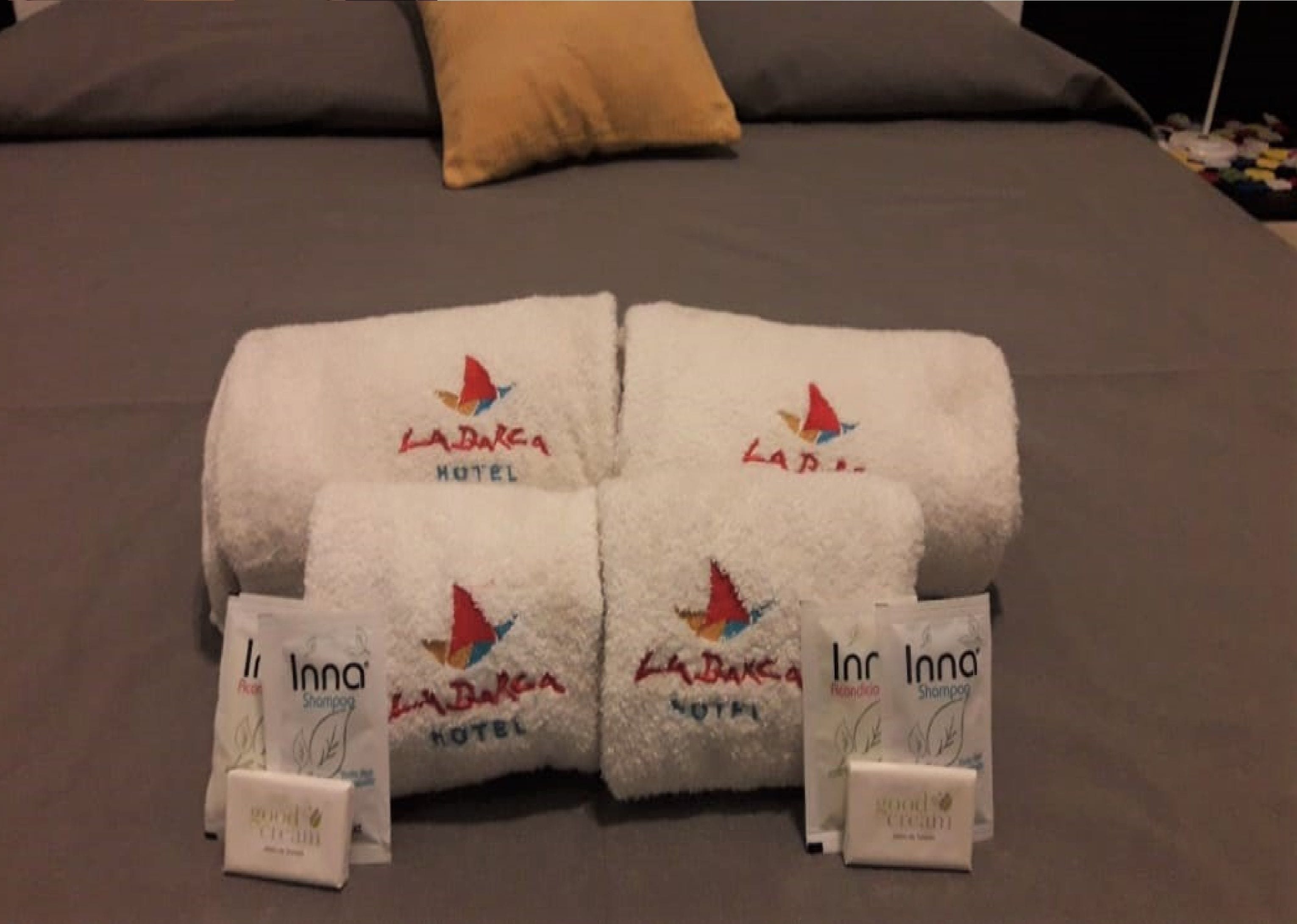 A bed with a grey headboard and a single yellow pillow. There are two white towels with the logo "La Barka Hotel" and two smaller towels with the logo "Inn Inn."