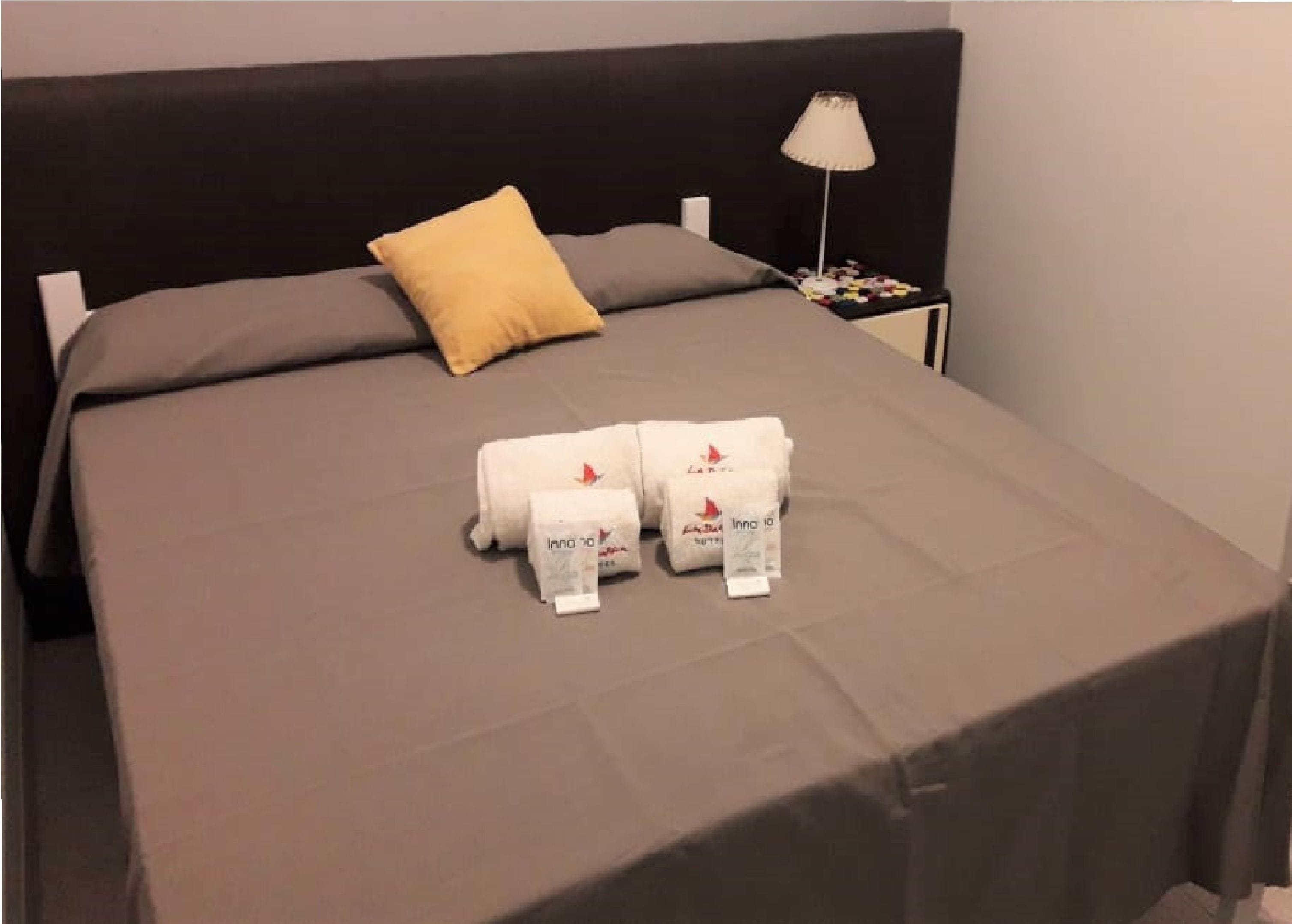 A neatly made bed with a grey bedspread, two white towels, and a single yellow pillow.