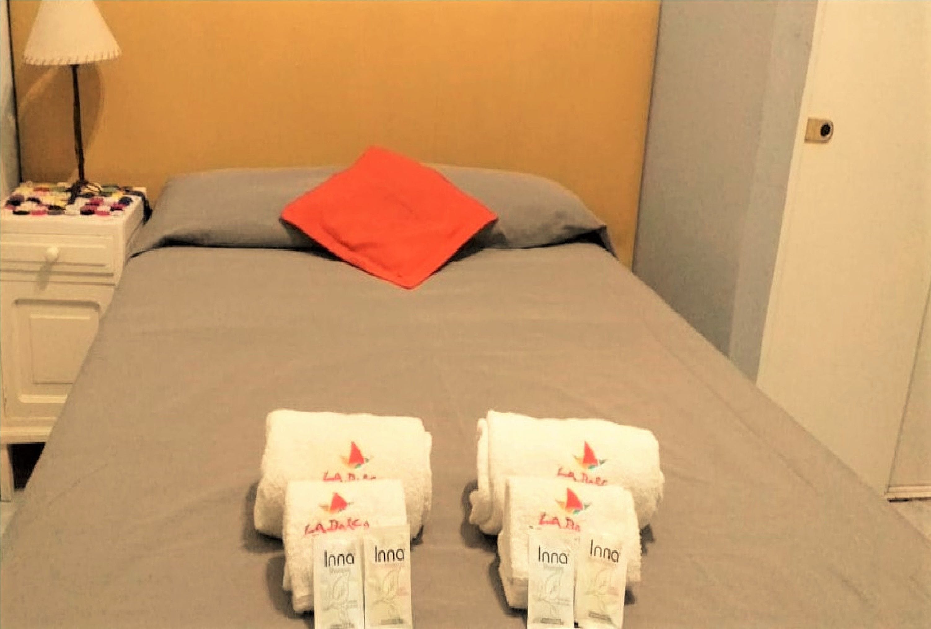 Een netjes opgemaakt bed met een grijze dekbedovertrek, een rood kussen en twee witte handdoeken met de merknaam 'La Bella'.