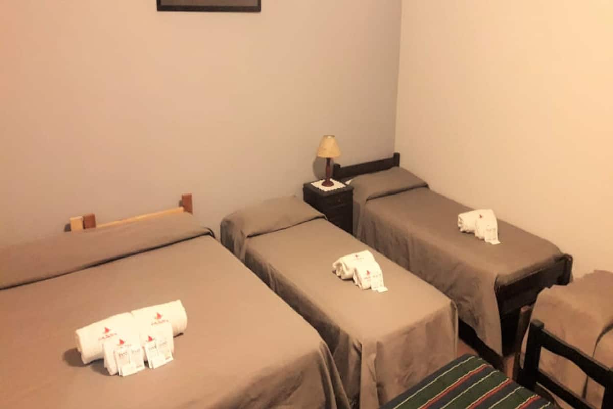 Habitación de hotel con dos camas individuales, cada una con una almohada y una toalla enrollada. Hay una mesita de noche con una lámpara entre las camas.