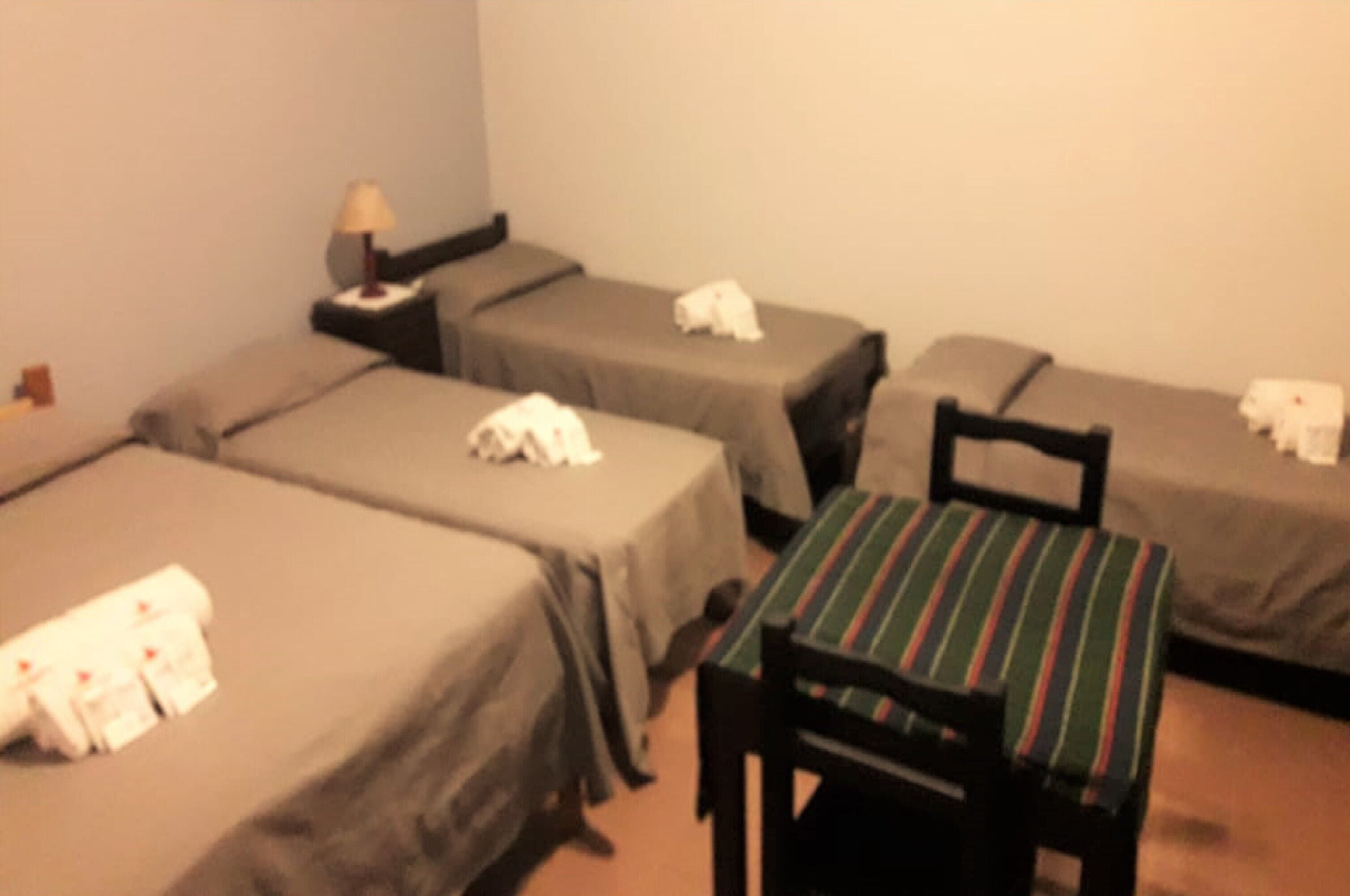 A hotel room with four single beds, a small table with a striped tablecloth, and a chair.