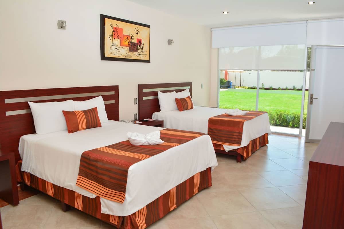 A hotel room with two beds, a painting on the wall, and a view of a green field through the door.