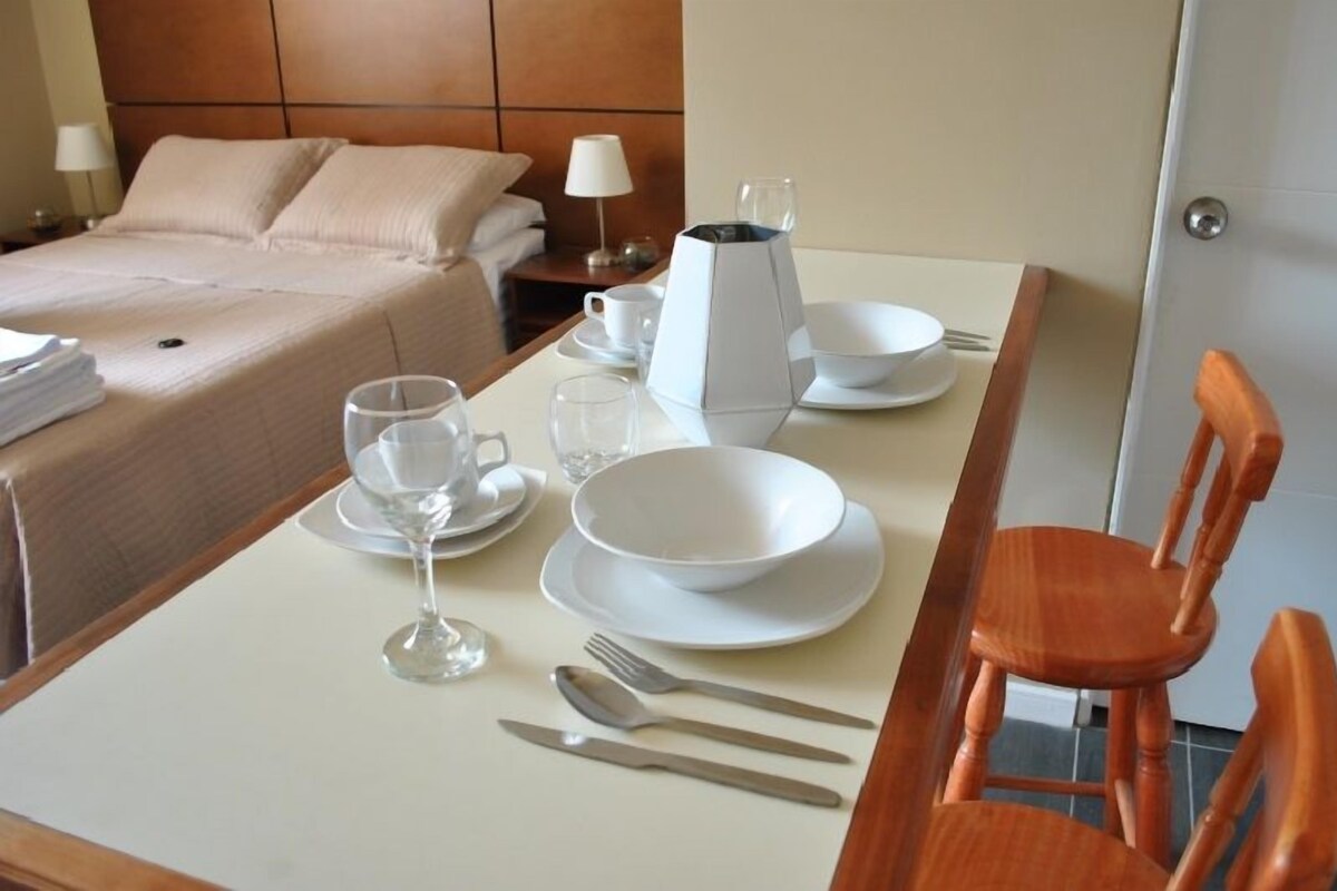 Una mesa de comedor bien dispuesta con platos, vasos, cubiertos y una cafetera en una habitación de hotel.