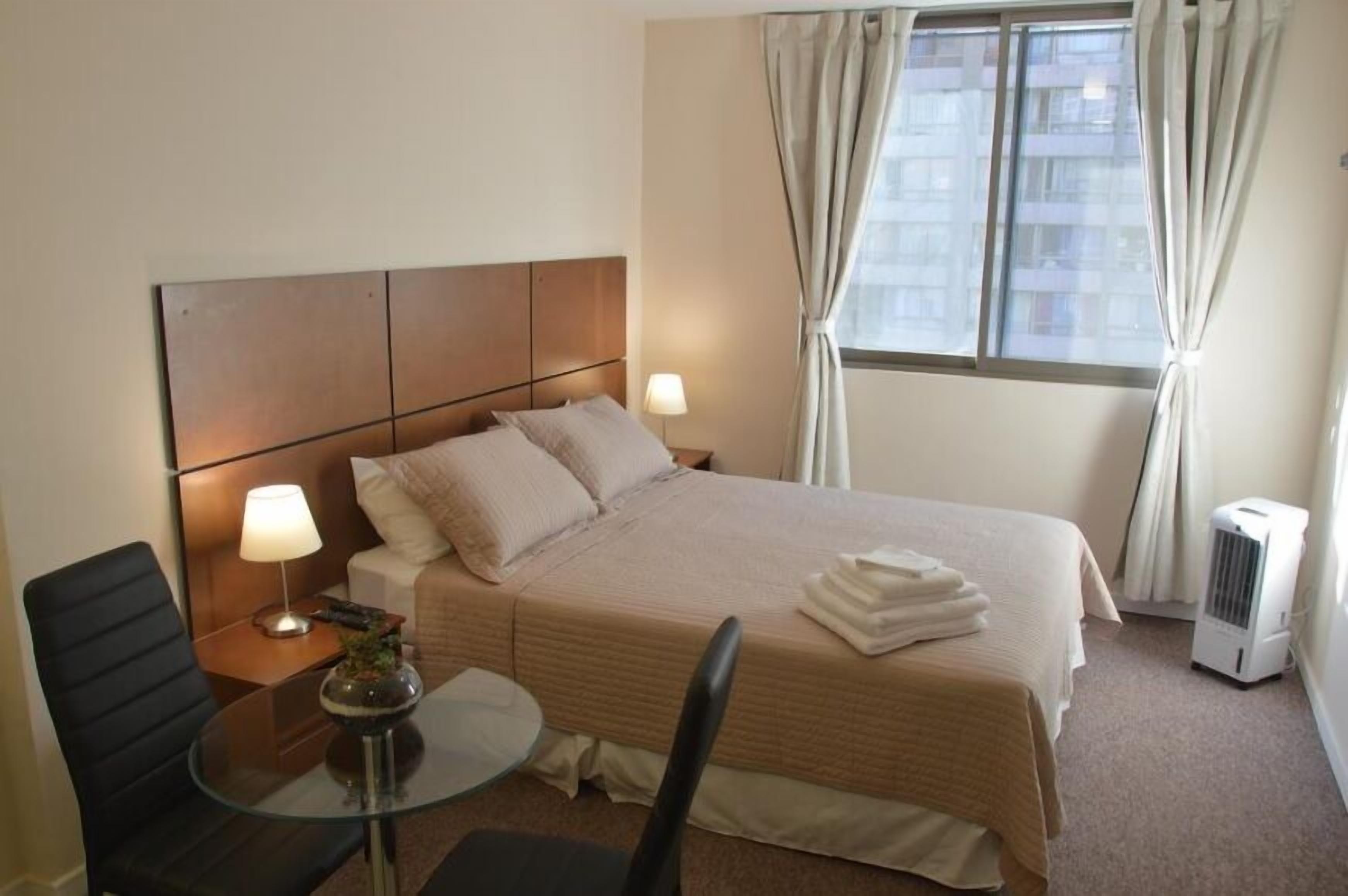Une chambre d’hôtel avec un lit, une table de chevet, une lampe, une chaise et une fenêtre avec des rideaux.