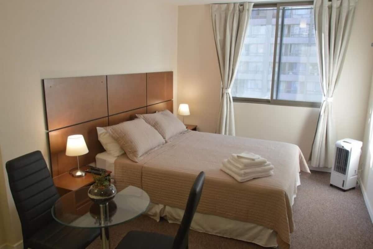 Une chambre d’hôtel avec un lit, une table de chevet, une lampe, une chaise et une fenêtre avec des rideaux.