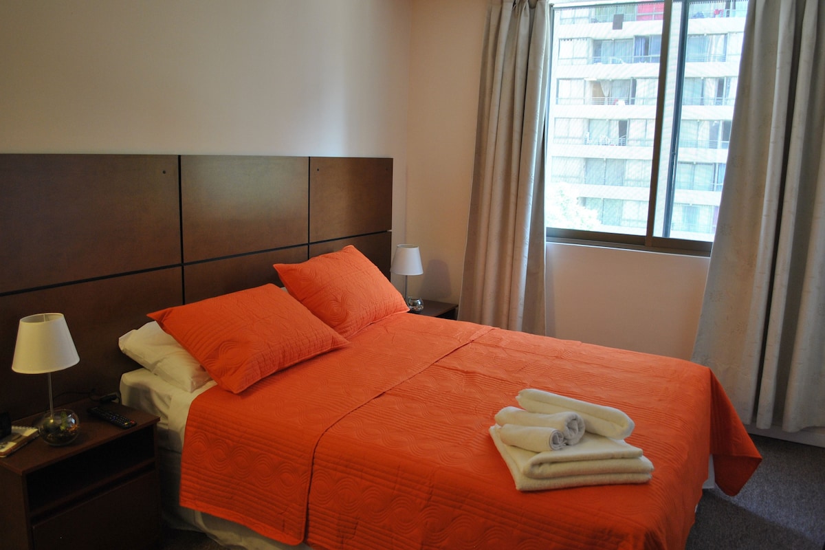 Habitación de hotel con cama, almohadas, mesita de noche, lámpara y ventana con cortinas.