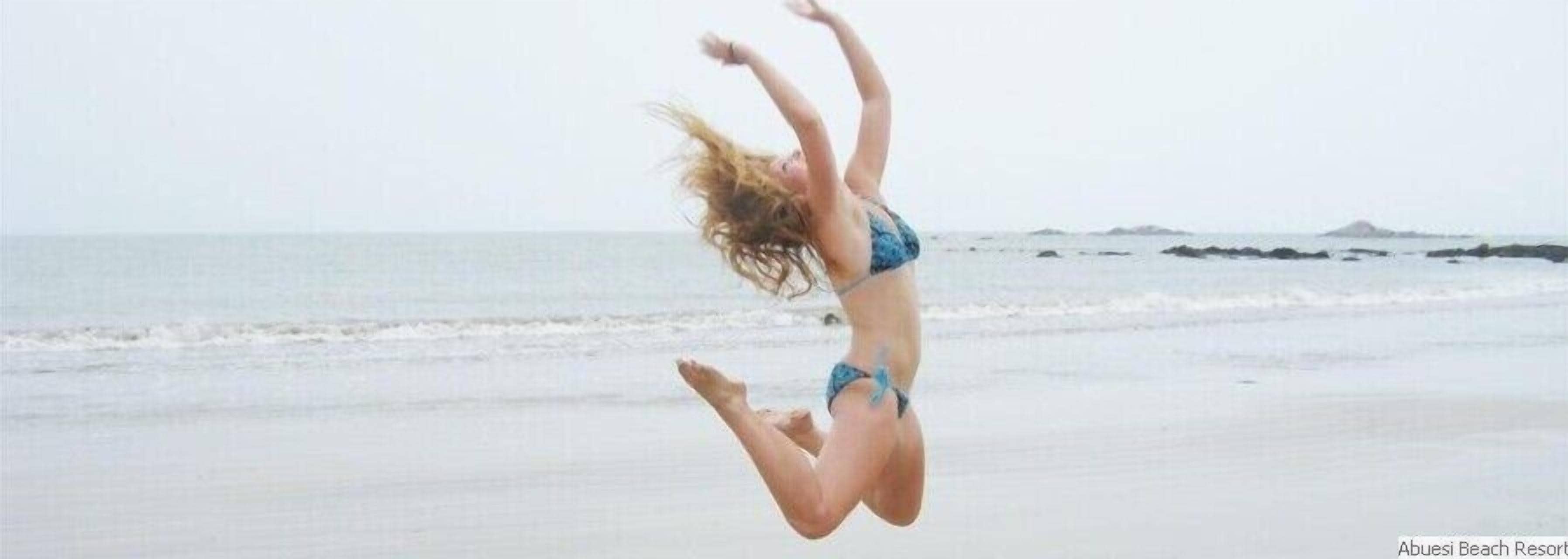 A person jumping on a beach with a clear view of the ocean and distant landforms.