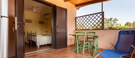 An open door leading to a bedroom with a bed, a table, and a chair. There is a balcony with a chair and a table.