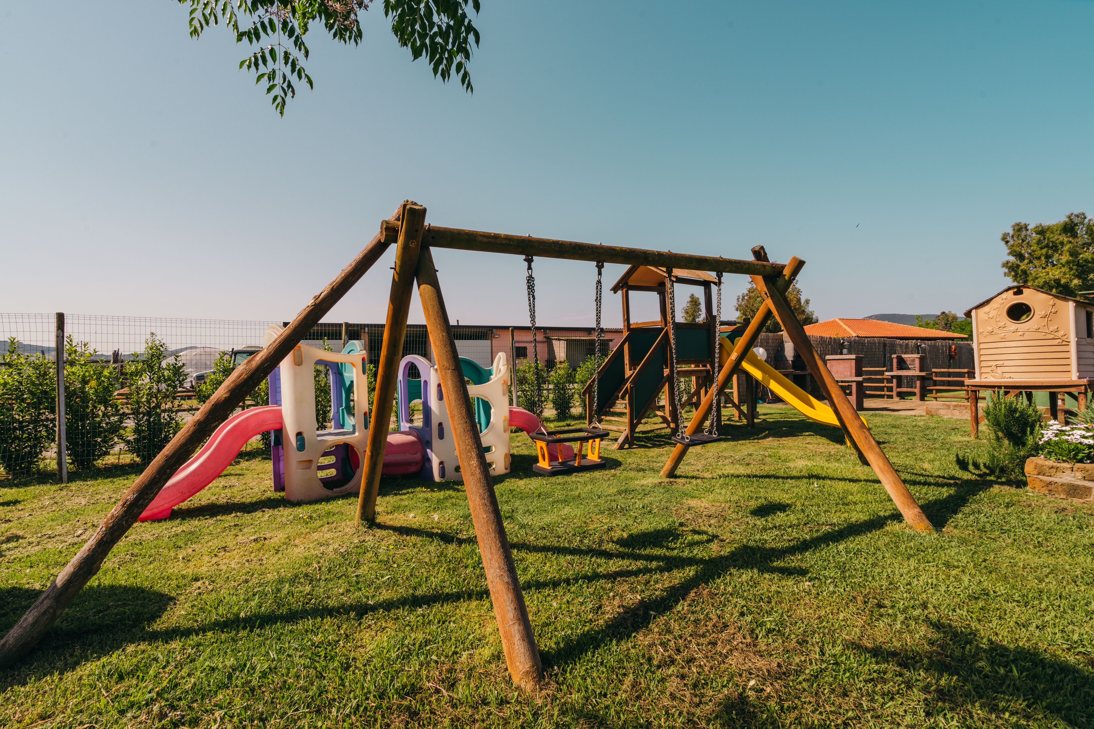 Un parque infantil con columpios y toboganes, rodeado de una cerca y edificios al fondo.