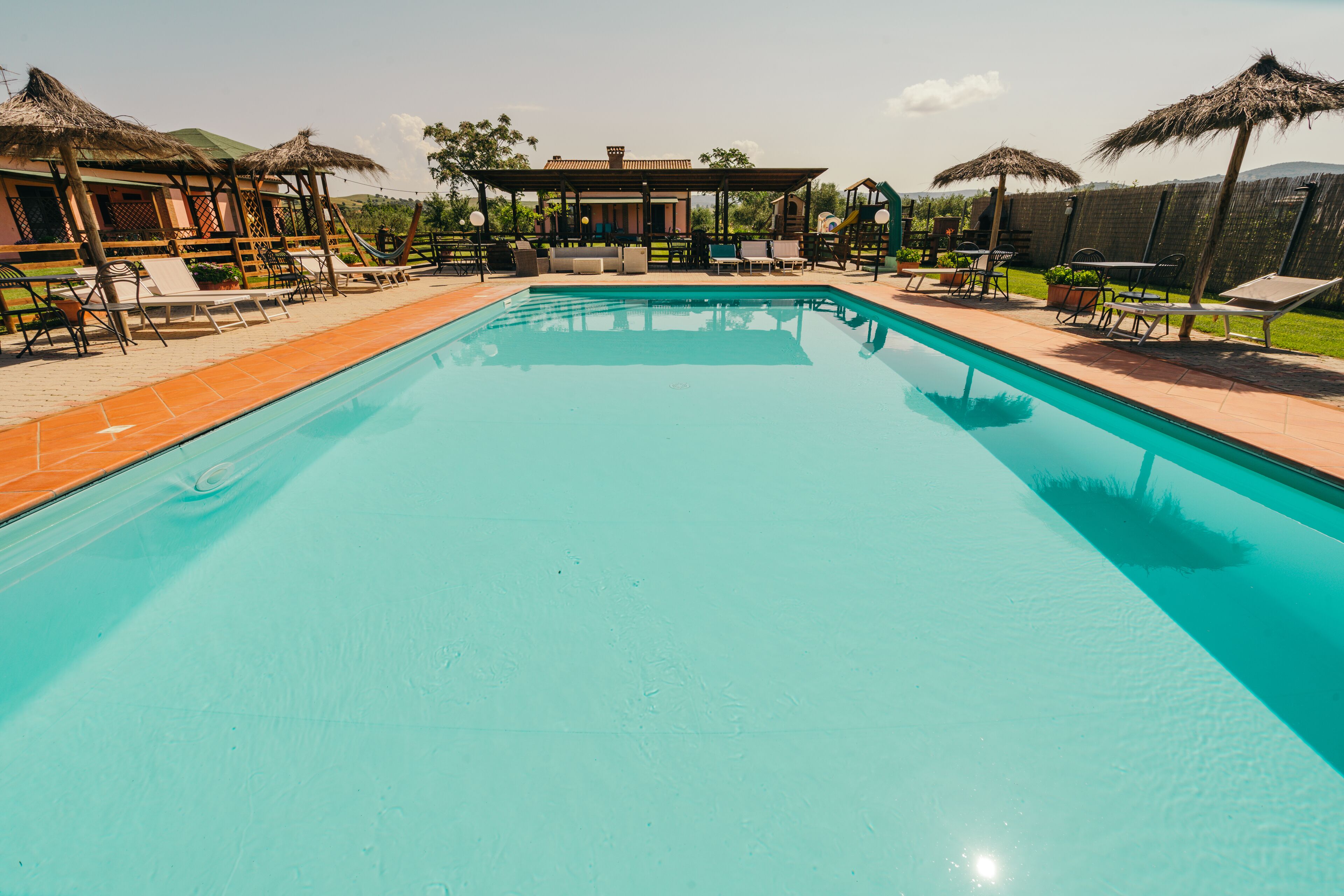 A swimming pool with lounge chairs and umbrellas, surrounded by a covered seating area and a fenced yard.