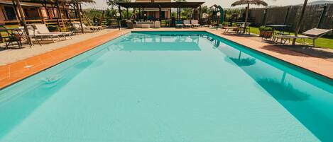 A swimming pool with lounge chairs and umbrellas, surrounded by a covered seating area and a fenced yard.