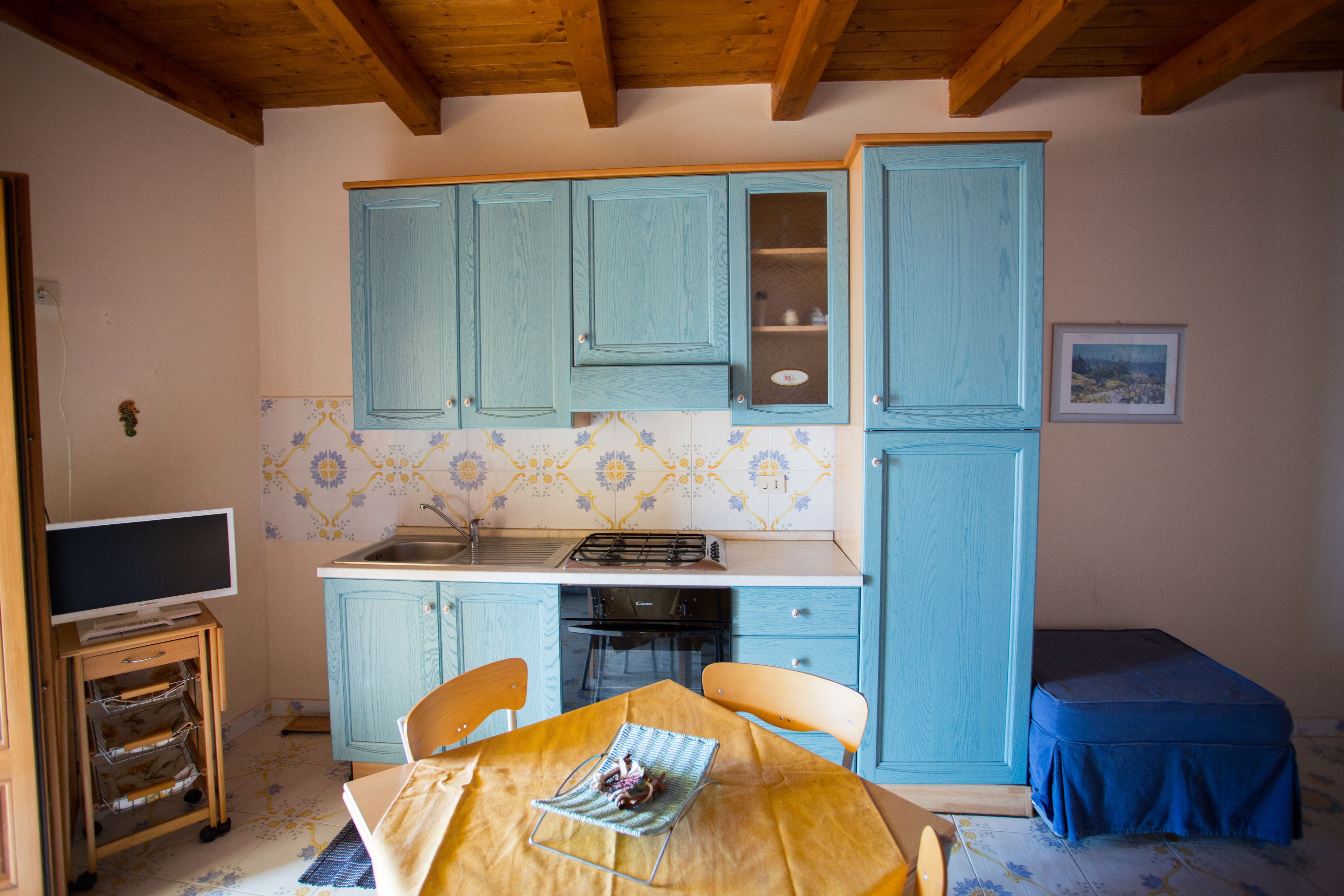 A compact kitchen with blue cabinets, a tiled backsplash, a dining table with chairs, and a small blue bench.