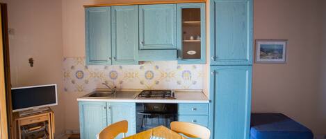 A compact kitchen with blue cabinets, a tiled backsplash, a dining table with chairs, and a small blue bench.