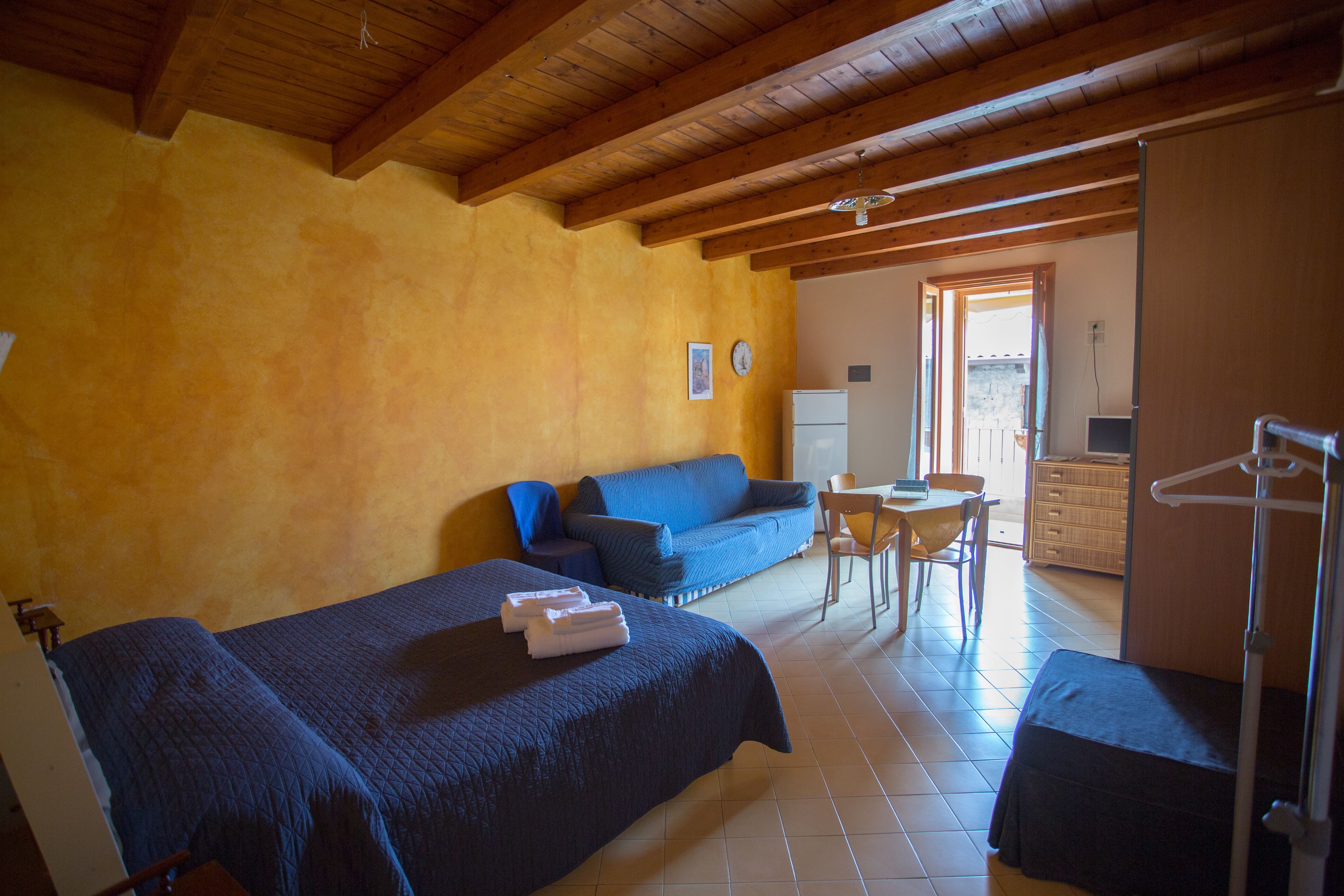Une pièce comprenant un lit, un canapé bleu, une petite table et une chaise. Le plafond est en bois et une porte donne accès à une autre pièce.