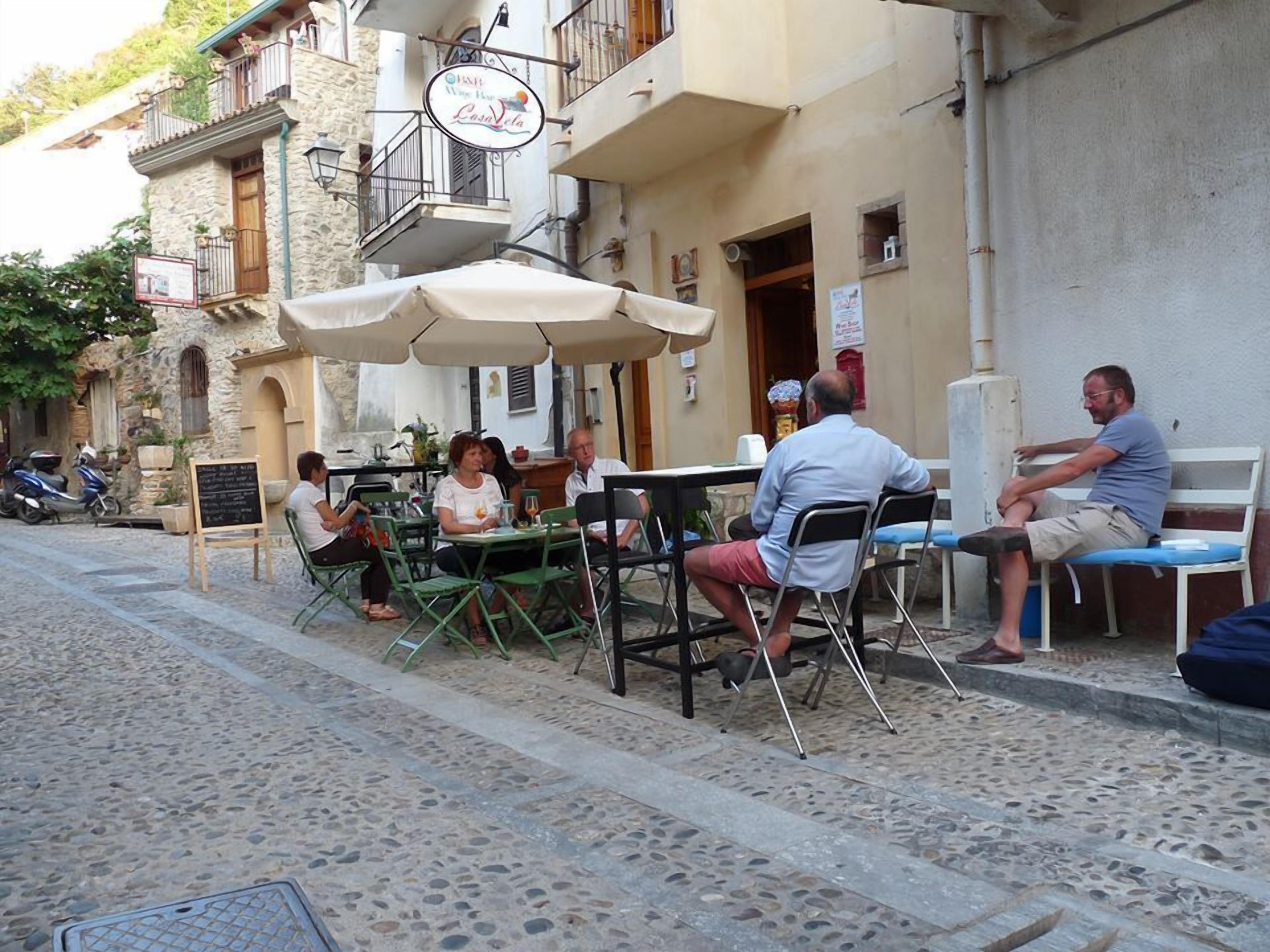 Un café al aire libre con mesas y sillas, un edificio de piedra con balcón y un letrero sobre la entrada.