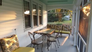 A covered porch with a glass table and metal chairs, a sofa with floral upholstery, and a view of a house and garden.