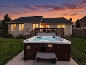 Une propriété résidentielle dotée d’un jacuzzi dans le jardin, d’une terrasse en bois et d’une maison dont les fenêtres sont illuminées.