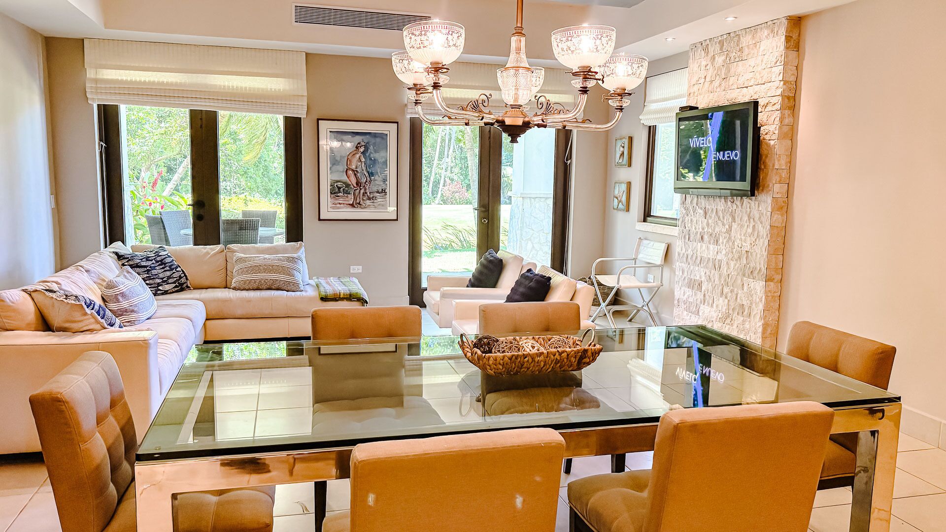 A modern living room with a glass dining table, two sofas, a chandelier, and a TV mounted on the wall.