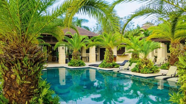 A pool area with palm trees, lounge chairs, and a building with a tiled roof.