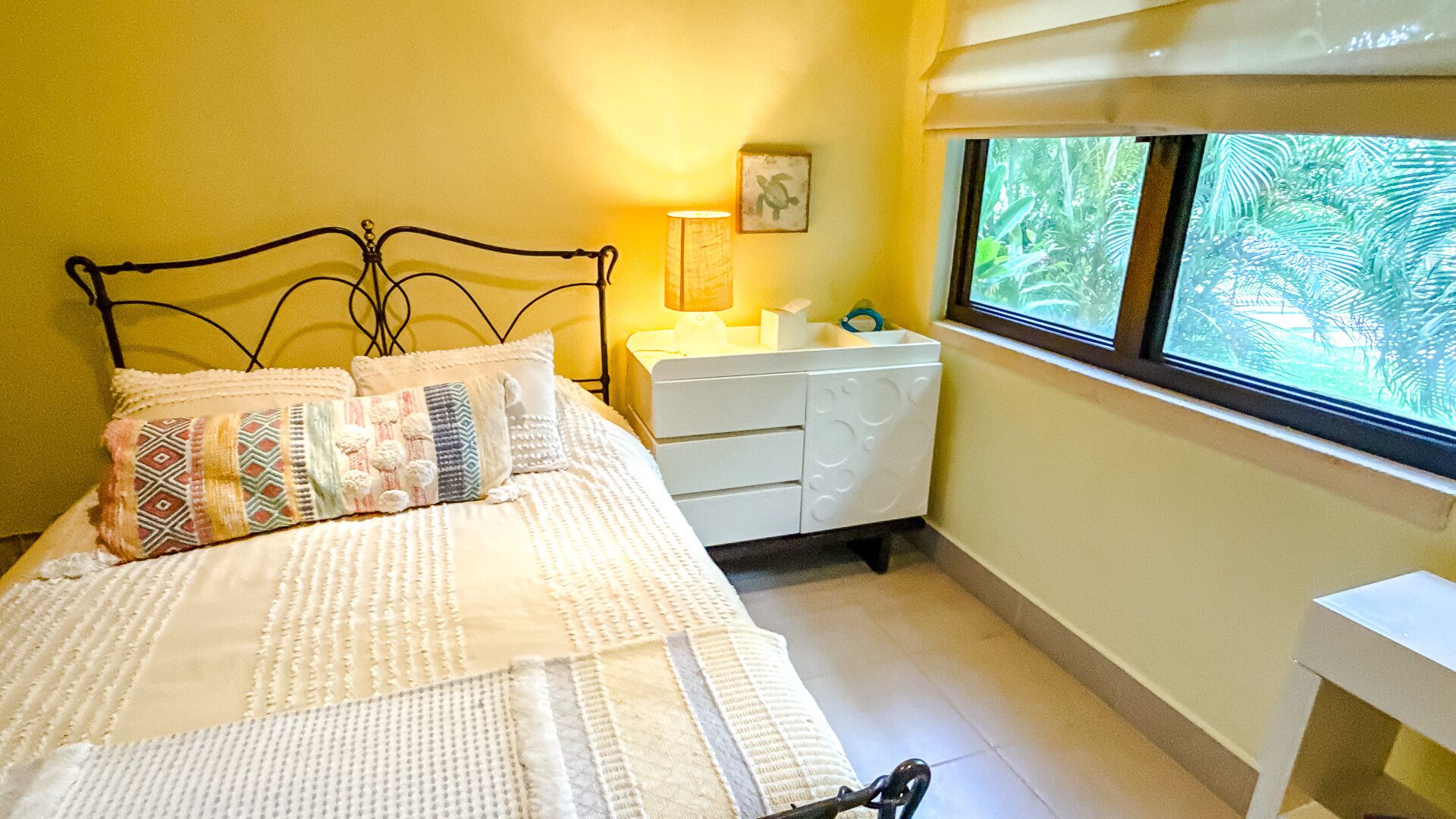 Una cama con ropa de cama estampada, una mesita de noche con lámpara, un pequeño marco de fotos y una ventana con vista a follaje.