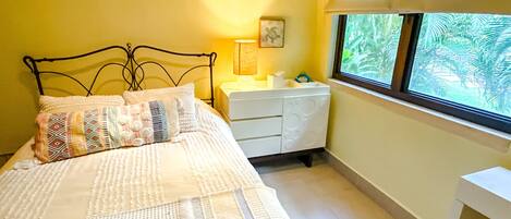 Una cama con ropa de cama estampada, una mesita de noche con lámpara, un pequeño marco de fotos y una ventana con vista a follaje.