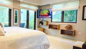 A bedroom with a large bed, a TV mounted on the wall, and a view of greenery through the windows.