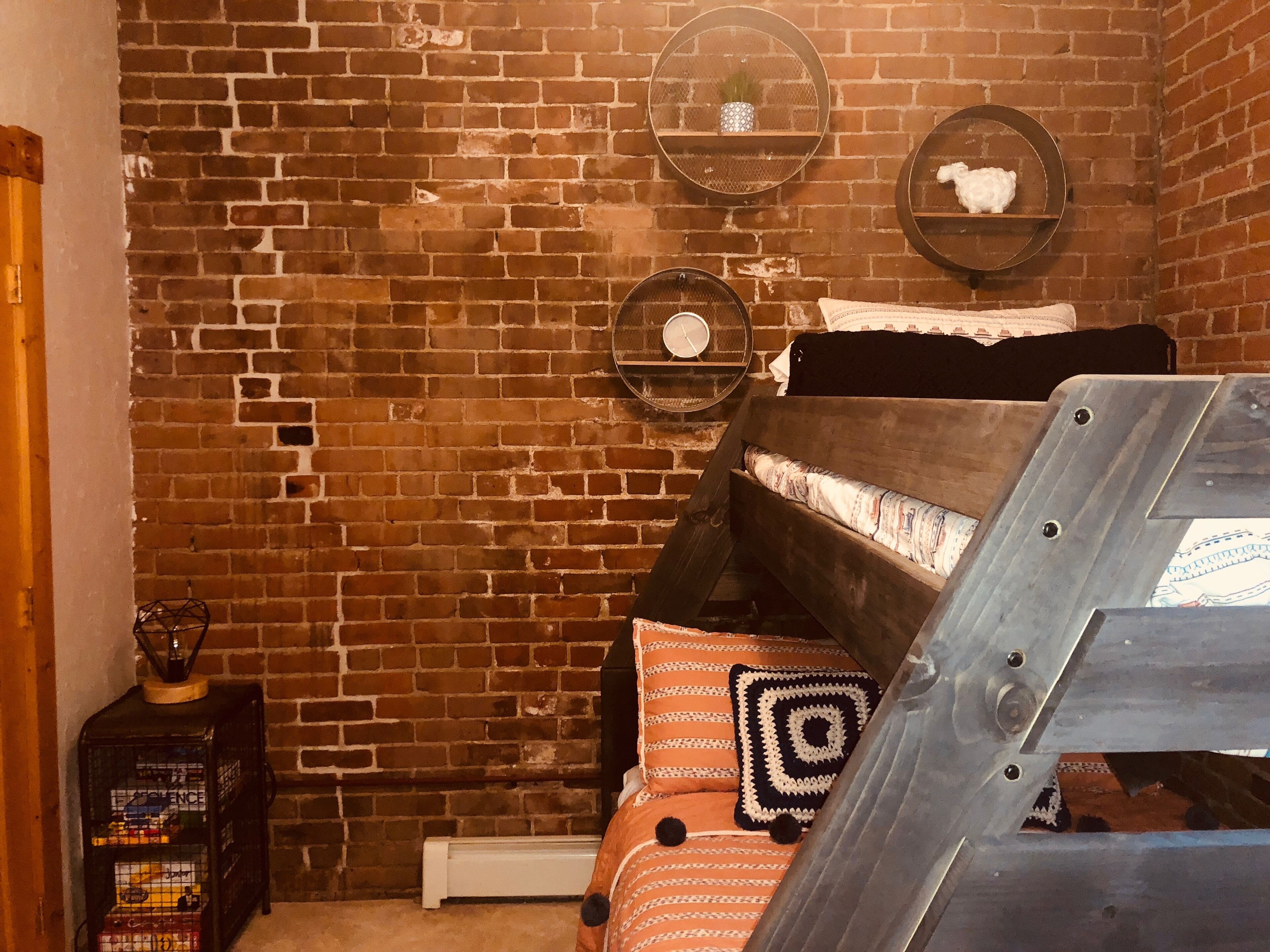 Ein Zimmer mit Backsteinwand, einem Bett mit gemusterten Betttextilien, zwei Wandregalen mit Dekoration und einem kleinen Tisch mit Lampe.
