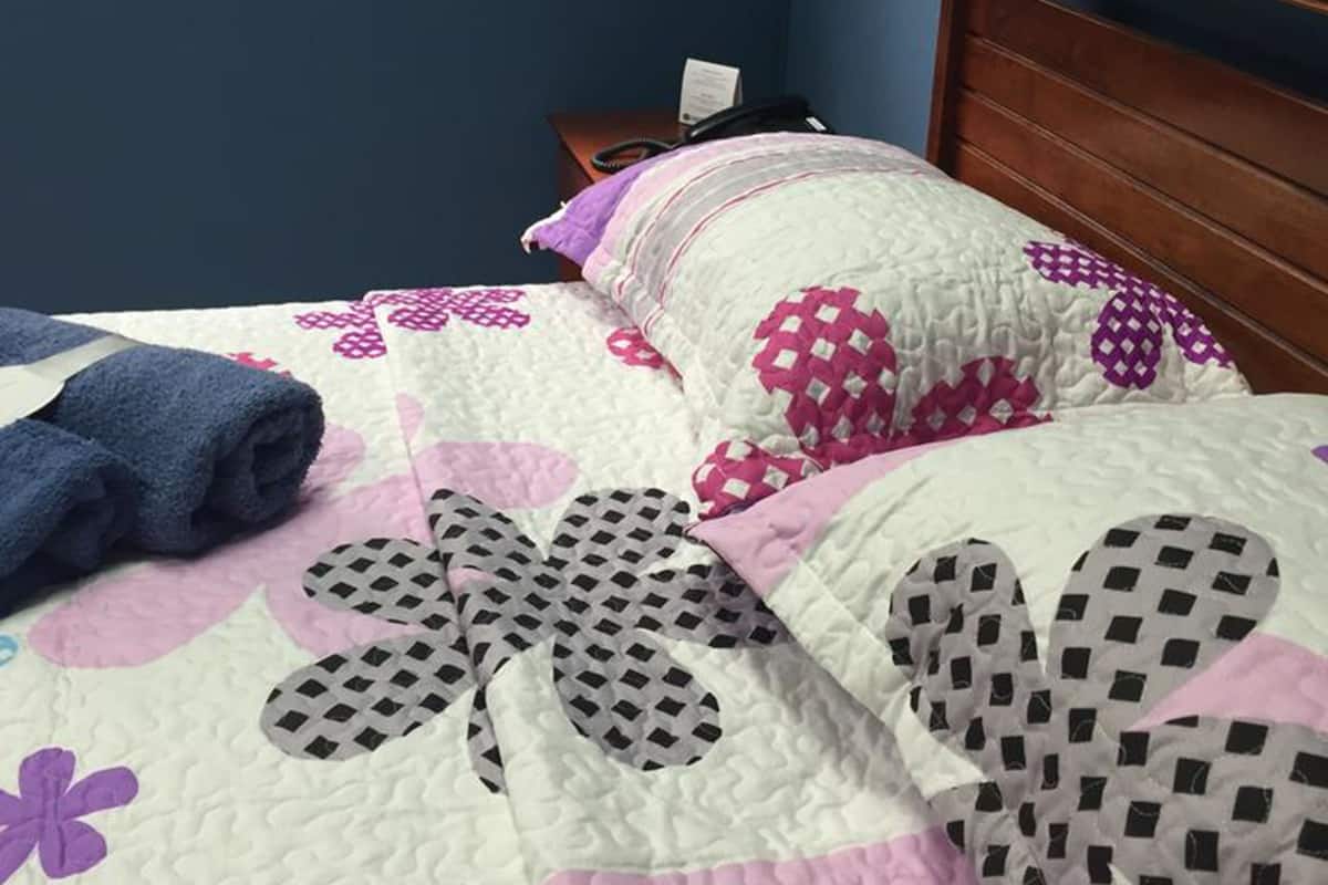 A bed with a floral-patterned quilt and pillows, a wooden headboard, a nightstand with a phone, and a window with a view of trees.