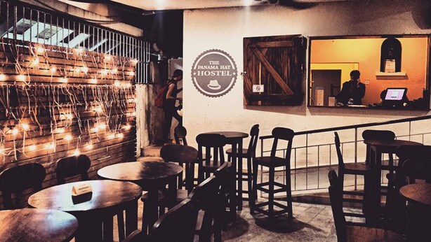 Un rincón acogedor de bar con mesas y sillas de madera, una escalera adornada con luces y un letrero del Hotel Panama Hat.