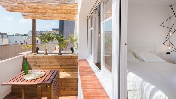Un balcon avec une table en bois, une bouteille et des verres, un lit avec une literie blanche et une vue sur la ville.