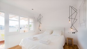 Une chambre à coucher au style minimaliste, avec un grand lit, du linge de lit blanc et des œuvres d’art géométriques accrochées au mur.