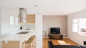 Une cuisine moderne dotée d’un îlot central, de tabourets de bar en bois, d’un téléviseur à écran plat et d’un canapé dans un espace de vie agrémenté de grandes fenêtres.