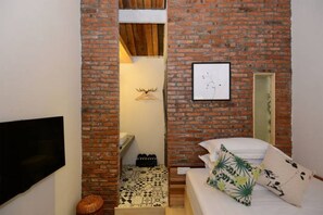 A compact room with a brick wall, a bed with patterned pillows, a mounted TV, and a small shelf with decorative items.