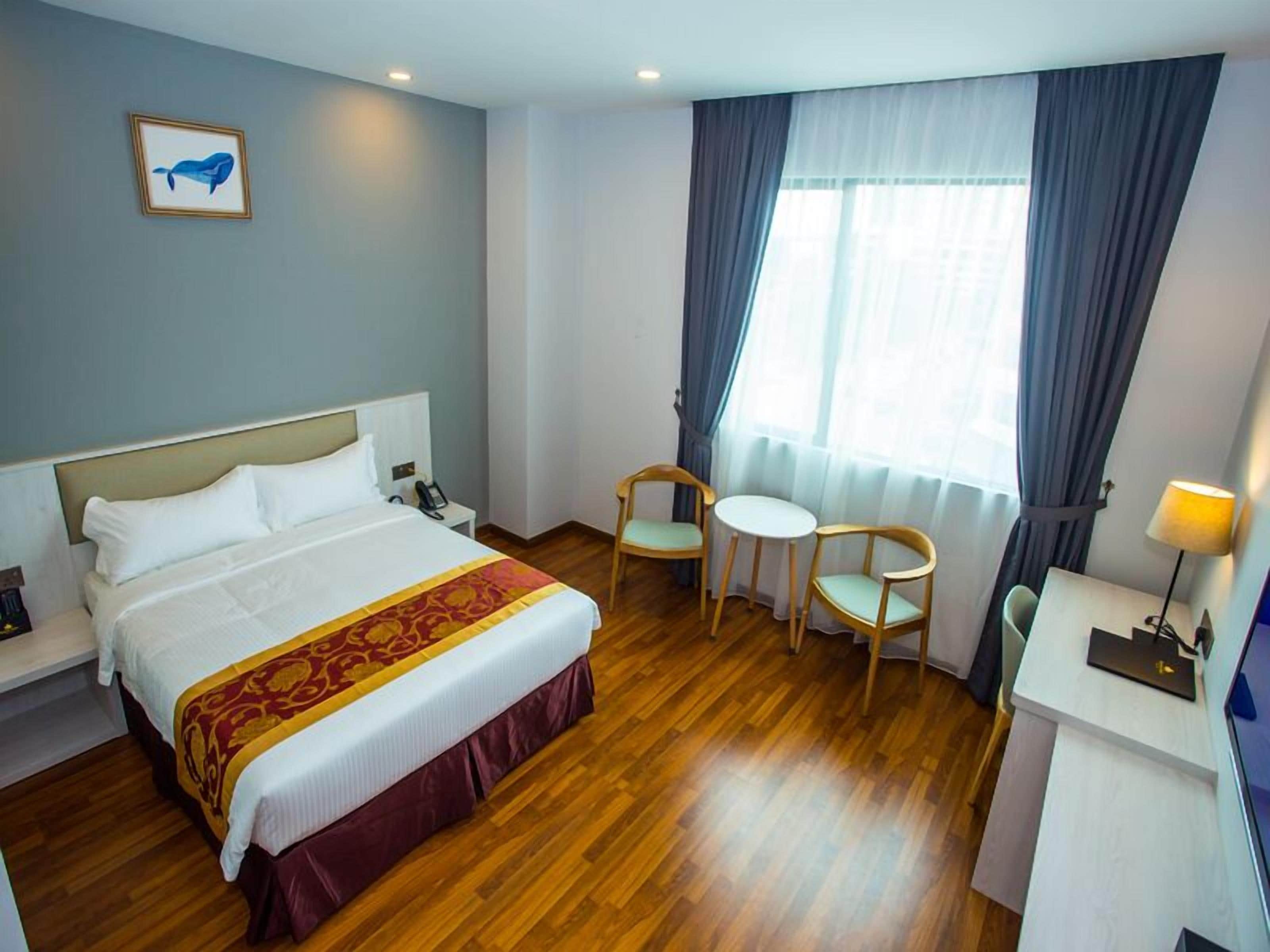 A hotel room with a bed, bedside table, lamp, two chairs, a small table, and a window with curtains.
