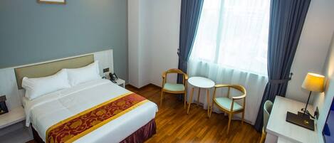 A hotel room with a bed, bedside table, lamp, two chairs, a small table, and a window with curtains.