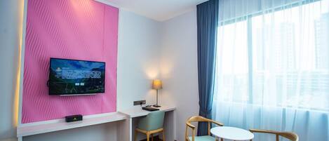 A hotel room with a pink accent wall, a flat-screen TV, a desk with a lamp, two chairs, and a large window with curtains.