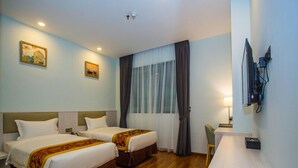 Ein Hotelzimmer mit zwei Betten, einem Schreibtisch, einem Stuhl, einem an der Wand montierten Fernseher und einem Fenster mit Vorhängen.